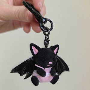 May include: A black plush bat keychain with pink accents on the ears, belly, and a small bib. The bat has large black wings and is attached to a black metal clip and ring.