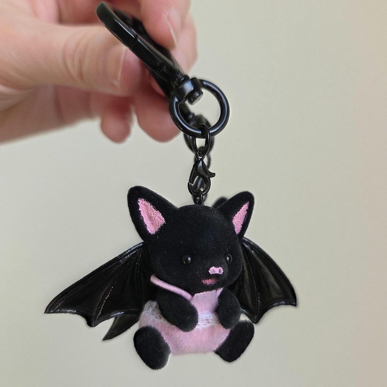 May include: A black plush bat keychain with pink accents on the ears, belly, and a small bib. The bat has large black wings and is attached to a black metal clip and ring.