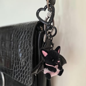 May include: A black bat-shaped keychain with pink accents, hanging from a black heart-shaped clasp. The bat has pink ears, a pink nose, and a pink and white dress. The keychain is attached to a black textured bag.
