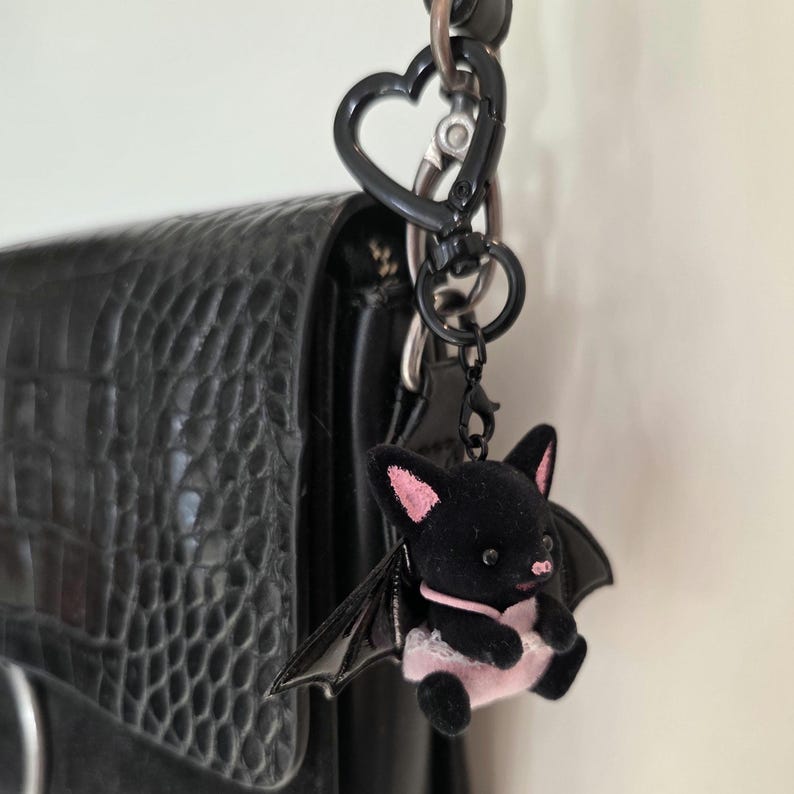 May include: A black bat-shaped keychain with pink accents, hanging from a black heart-shaped clasp. The bat has pink ears, a pink nose, and a pink and white dress. The keychain is attached to a black textured bag.
