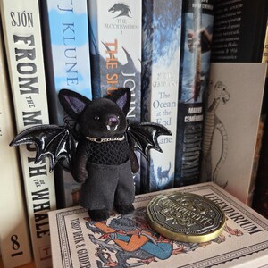 Puede incluir: Una mu&ntilde;eca de murci&eacute;lago de peluche negro con alas grandes y brillantes, que lleva un peque&ntilde;o collar de cadena dorada y pantalones grises. El murci&eacute;lago est&aacute; de pie sobre una baraja de cartas del tarot y est&aacute; rodeado de libros. Tambi&eacute;n se ve una moneda dorada con texto.