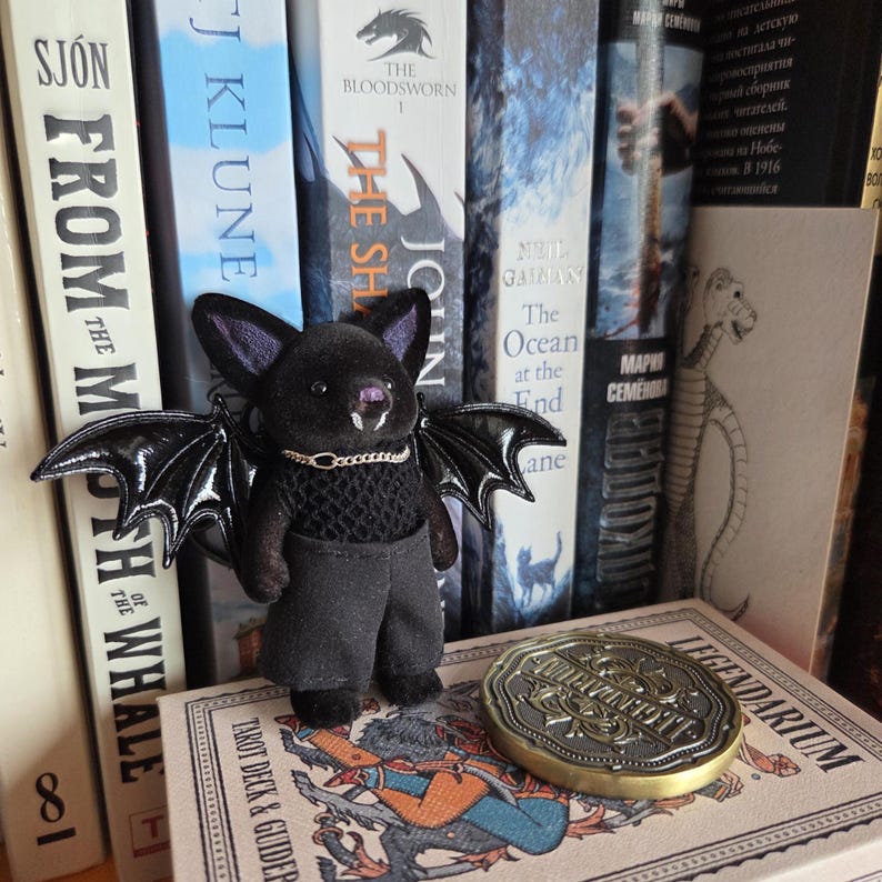 Puede incluir: Una mu&ntilde;eca de murci&eacute;lago de peluche negro con alas grandes y brillantes, que lleva un peque&ntilde;o collar de cadena dorada y pantalones grises. El murci&eacute;lago est&aacute; de pie sobre una baraja de cartas del tarot y est&aacute; rodeado de libros. Tambi&eacute;n se ve una moneda dorada con texto.