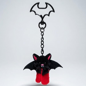 May include: A black bat-shaped keychain with a chain and a small plush bat. The plush bat has black wings, ears, and body, with red accents on the ears and feet. The keychain is suspended against a white background.