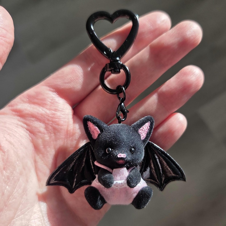 May include: A black bat keychain with pink accents. The plush bat has black wings, pink ears, and a pink belly. The keychain has a black heart-shaped clasp.