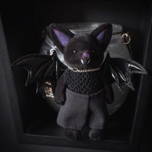 Puede incluir: Una figura de murci&eacute;lago negro de peluche con orejas internas moradas, un peque&ntilde;o collar de cadena dorada y alas negras brillantes. El murci&eacute;lago lleva un top de encaje negro y pantalones cortos negros. La figura est&aacute; en un entorno oscuro y sombreado.