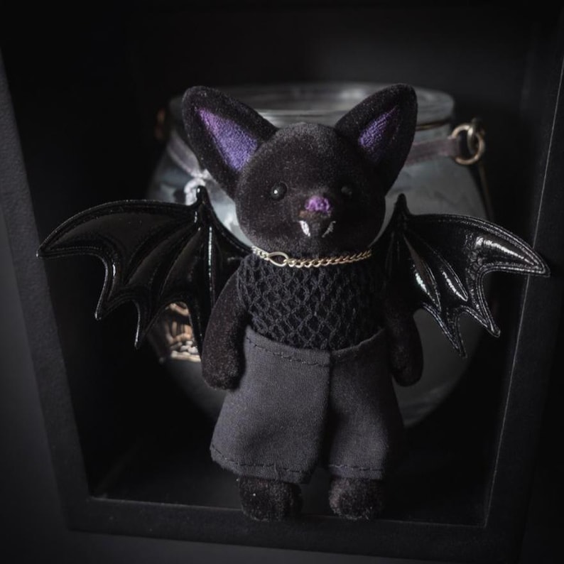 Puede incluir: Una figura de murci&eacute;lago negro de peluche con orejas internas moradas, un peque&ntilde;o collar de cadena dorada y alas negras brillantes. El murci&eacute;lago lleva un top de encaje negro y pantalones cortos negros. La figura est&aacute; en un entorno oscuro y sombreado.