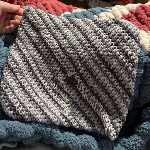 May include: A handmade, square crochet item in shades of gray and purple. The stitches are visible, creating a textured surface. The item is resting on a blue, red, and white blanket.