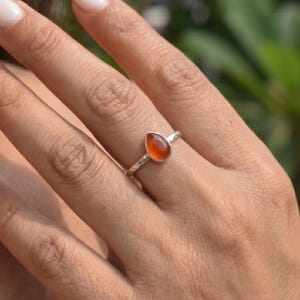 May include: A silver ring featuring a teardrop-shaped orange carnelian gemstone set in a smooth bezel. The ring band has a textured, hammered finish that catches the light, providing a subtle contrast to the polished surface of the stone.