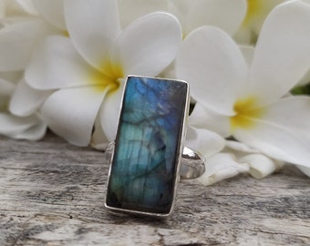May include: A rectangular silver ring featuring a labradorite stone with blue and green iridescent colors. The ring is set against a backdrop of white flowers and a weathered wooden surface, creating a natural aesthetic.