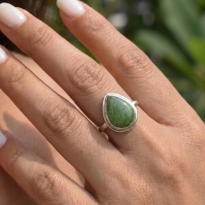 May include: A silver ring featuring a teardrop-shaped green stone set in a smooth metal bezel. The stone displays natural mottled textures in various shades of forest and olive green, securely mounted on a simple, polished silver band.