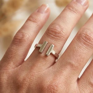 May include: A silver-toned open ring featuring two T-shaped ends that face each other with a gap in the centre. The band has a smooth, polished metallic finish and a simple, clean design that wraps partially around the finger.