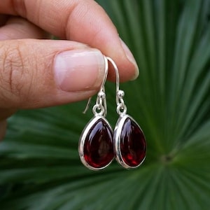 May include: A pair of silver drop earrings featuring smooth, teardrop-shaped dark red stones set in polished metal bezels. The earrings hang from simple silver hooks, showcasing a deep crimson hue and a reflective, glossy surface finish.