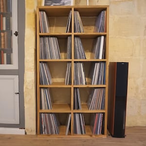 Peut inclure: Une étagère à disques en bois avec 12 compartiments remplis de disques vinyles. L'étagère est placée devant un mur en pierre et à côté d'une enceinte noire.
