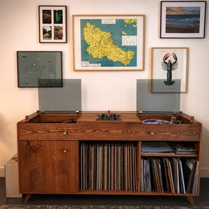 Può includere: Una console multimediale in legno dotata di due giradischi integrati con coperchi antipolvere grigi sulla superficie superiore. L'unità include un'anta a sinistra, uno scomparto verticale per dischi in vinile al centro e ripiani a destra.