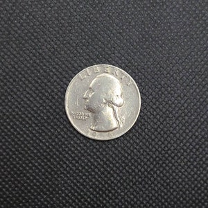 May include: A silver US quarter coin with the words "Liberty" and "In God We Trust" on the front. The year 1965 is visible on the coin.
