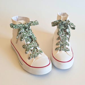 Puede incluir: Par de zapatillas altas blancas con cordones estampados florales verdes. Las zapatillas tienen una franja roja alrededor de la suela y ojales metálicos. Los cordones están atados con lazos.