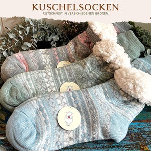 Könnte beinhalten: Drei Paar Kuschelsocken mit Fair-Isle-Muster in Blau-, Grau- und Rosatönen. Jede Socke hat ein weißes Sherpa-Fleece-Futter am Bündchen und ein rundes Etikett mit einer Schaf-Illustration. Der Text KUSCHELSOCKEN RUTSCHFEST IN VERSCHIEDENEN GRÖSSEN steht am oberen Bildrand.