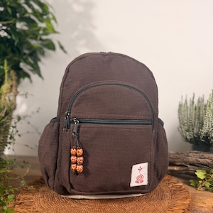 Könnte beinhalten: Ein dunkelbrauner Rucksack mit strukturiertem Fischgrätenmuster, ausgestattet mit einer vorderen Reißverschlusstasche, an deren Zipper drei Holzperlen befestigt sind, sowie einem kleinen weißen Aufnäher mit roter Spiral- und Sprossgrafik.