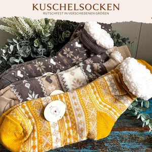 Könnte beinhalten: Drei Paar Strickstrümpfe mit einem weiß-braun-senfgelben Muster im nordischen Stil. Jeder Strumpf hat einen weißen, flauschigen Sherpa-Umschlag am oberen Rand und ein rundes Etikett mit einem Schaf-Motiv. Der Text KUSCHELSOCKEN ist zu sehen.
