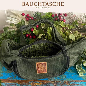 Könnte beinhalten: Eine dunkelgrüne Bauchtasche aus Cord mit schwarzem Reißverschluss, verstellbarem Riemen und einem quadratischen Aufnäher mit goldfarbenem Grafikdesign auf der Vorderseite. Das Innenfutter ist hellgrün mit einem schwarzen geometrischen Muster.