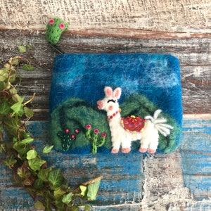 Könnte beinhalten: Eine blaue Filztasche mit einem skurrilen Lama-Design. Das weiße Lama hat rosa Ohren und Füße, einen roten und orangefarbenen Sattel und ist vor dem Hintergrund grüner Hügel und eines blauen Himmels platziert. Ein grüner Kaktus und rosa Blumen sind ebenfalls abgebildet.