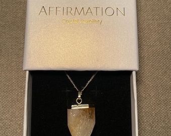Puede incluir: Un collar de cadena dorada con un colgante de cristal de cuarzo transparente en forma de punta. El collar está en una caja blanca con las palabras "AFFIRMATION Crystal Jewellery" impresas en la parte superior.