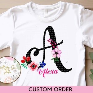 May include: White t-shirt featuring a black letter A adorned with pink, red, and blue flowers. The name "Alexa" is written in pink beneath the letter.