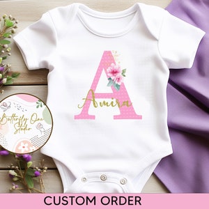 May include: A white baby onesie with a pink letter A decorated with flowers and gold polka dots. The name Amira is written in gold script below the letter. The onesie is on a wooden surface with a pink and green logo in the background.