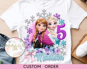 Puede incluir: Camiseta blanca con un diseño personalizado que presenta a los personajes Elsa y Anna de Frozen. El diseño incluye copos de nieve, flores y el número 5. El nombre Madison está escrito en azul en la camiseta.  PEDIDO PERSONALIZADO.