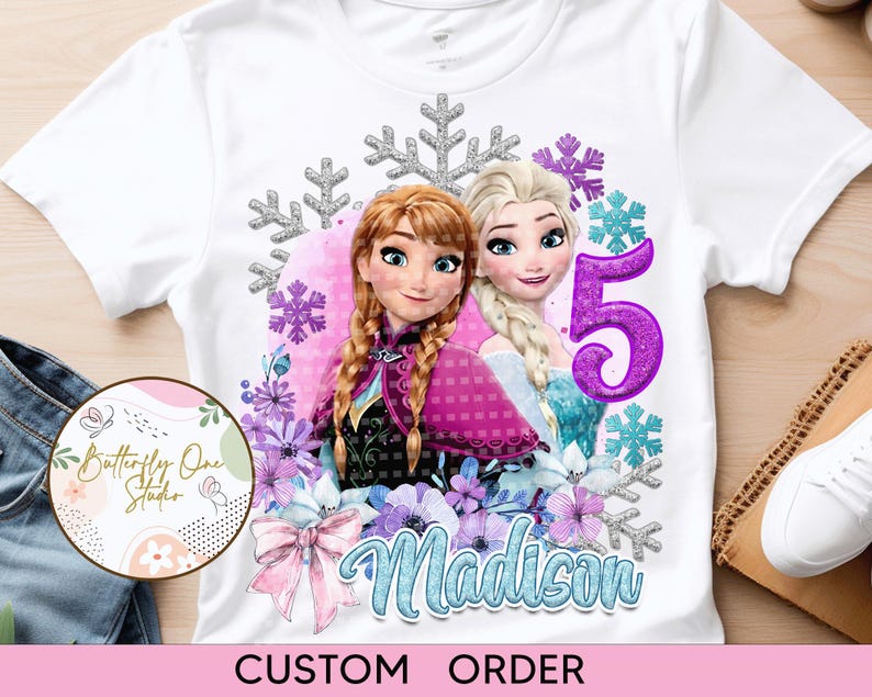 May include: White t-shirt with a custom design featuring the characters Elsa and Anna from Frozen. The design includes snowflakes, flowers, and the number 5. The name Madison is written in blue on the shirt.  CUSTOM ORDER.