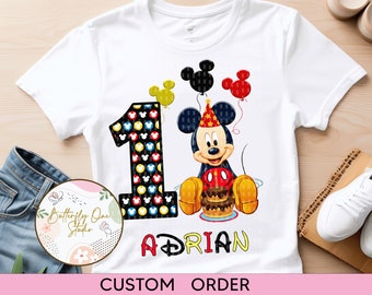 Op de afbeelding: Wit T-shirt met een zwart en geel nummer één, Mickey Mouse die een verjaardagstaart vasthoudt, en de naam "Adrian" in gele en zwarte letters. Het shirt heeft onderaan de tekst "Klantorder".
