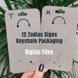 May include: Two grey cardstock packaging templates for keychains featuring the zodiac signs Aries and Pisces. The text "12 Zodiac Signs Keychain Packaging" and "Digital Files" is printed on the cards.