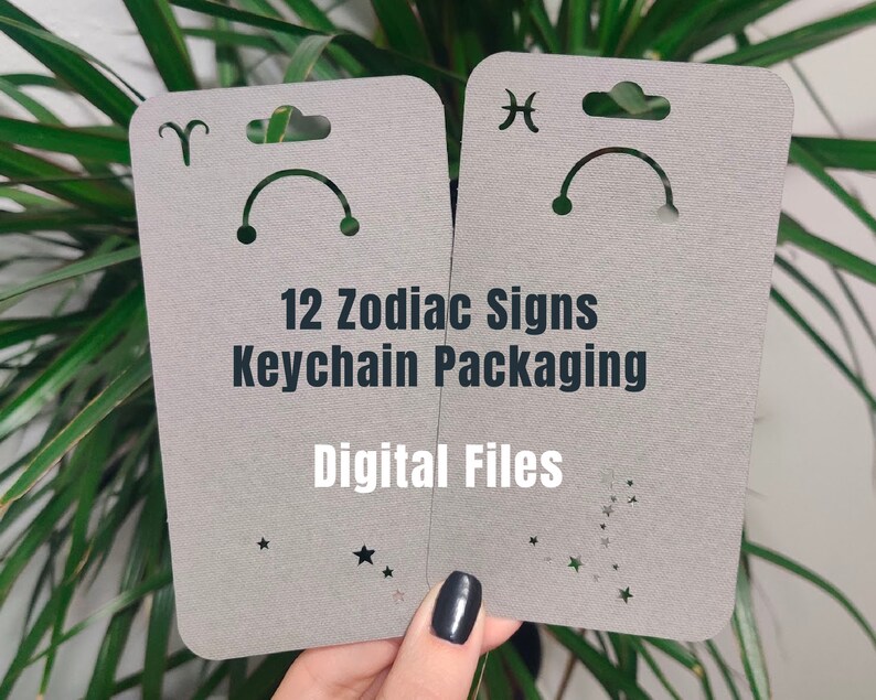 May include: Two grey cardstock packaging templates for keychains featuring the zodiac signs Aries and Pisces. The text "12 Zodiac Signs Keychain Packaging" and "Digital Files" is printed on the cards.