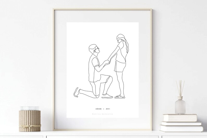 May include: Framed minimalist line art print depicting a marriage proposal. A man kneels before a woman, holding her hands. The artwork is in black and white, with the names "ADAM + ANI" at the bottom. The print is displayed in a white frame.