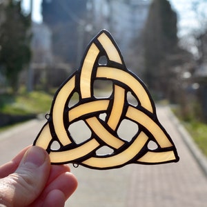 May include: A stained glass Celtic knot, also known as a Trinity knot, in the shape of a triangle. The knot is made of yellow glass with black outlines. The knot is held in a hand against a blurred background.