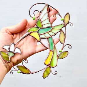 May include: A stained glass hummingbird with green, yellow, and white glass pieces. The hummingbird is perched on a vine with green leaves and beads.