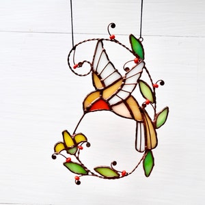 May include: A stained glass hummingbird with yellow, red, and white wings, feeding from a yellow flower. The hummingbird is suspended from a wire and surrounded by green leaves and red berries.