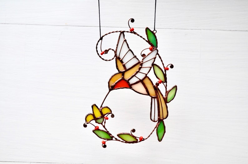 May include: A stained glass hummingbird with yellow, red, and white wings, feeding from a yellow flower. The hummingbird is suspended from a wire and surrounded by green leaves and red berries.