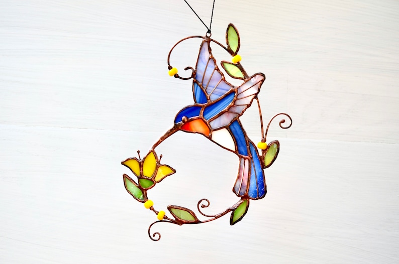 May include: A stained glass hummingbird with blue, pink, and orange wings, feeding from a yellow flower. The hummingbird is suspended from a wire with yellow beads and green leaves.