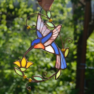 May include: A stained glass hummingbird with blue, pink, and orange wings, feeding from a yellow flower. The hummingbird is suspended from a wire and surrounded by green leaves.