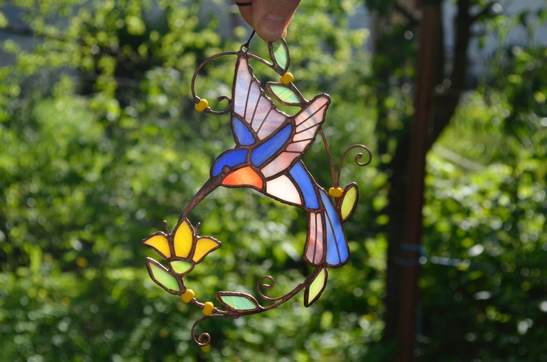 May include: A stained glass hummingbird with blue, pink, and orange wings, feeding from a yellow flower. The hummingbird is suspended from a wire and surrounded by green leaves.
