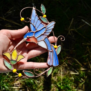 May include: A stained glass hummingbird with blue, white, and red wings, perched on a copper wire stem with yellow flowers and green leaves.