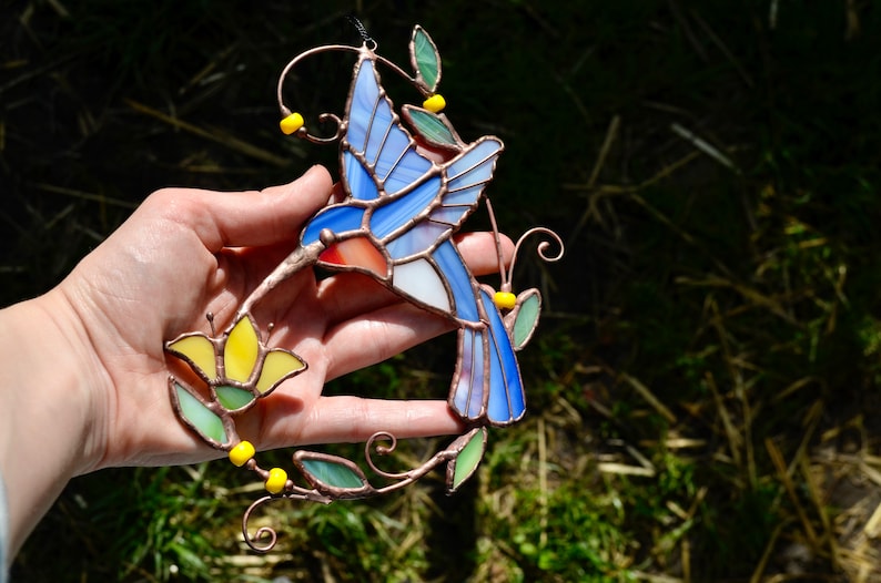 May include: A stained glass hummingbird with blue, white, and red wings, perched on a copper wire stem with yellow flowers and green leaves.