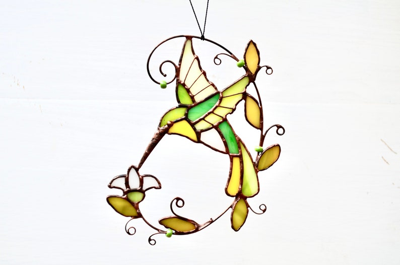 May include: A stained glass hummingbird with yellow, green, and white accents, perched on a branch with green leaves and beads. The hummingbird is in flight, with its beak extended towards a flower.