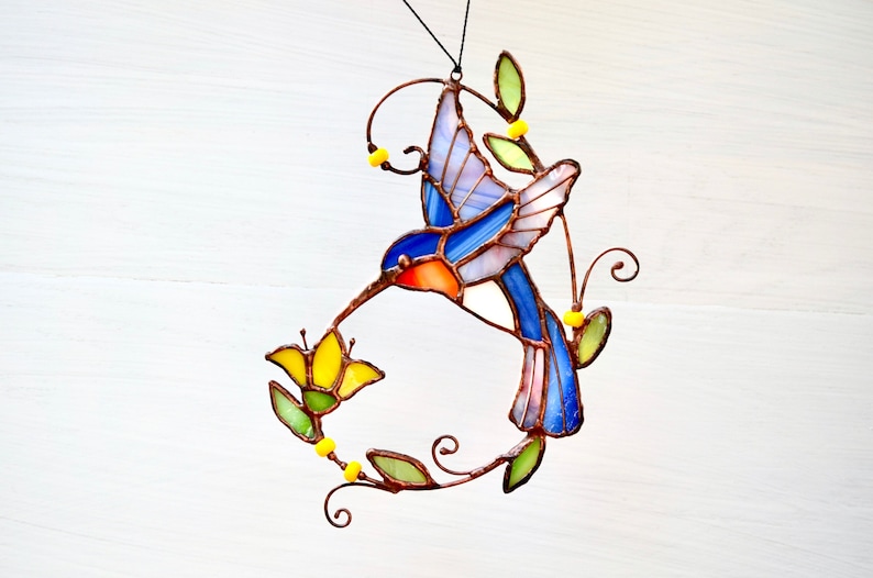 Pu&ograve; includere: Un colibr&igrave; in vetro colorato con ali blu, rosa e arancioni, che si nutre da un fiore giallo. Il colibr&igrave; &egrave; sospeso da un filo di rame con perline gialle.
