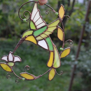 May include: A stained glass hummingbird with green, yellow, and white glass pieces, perched on a branch with a white flower. The hummingbird is in flight with its wings spread.