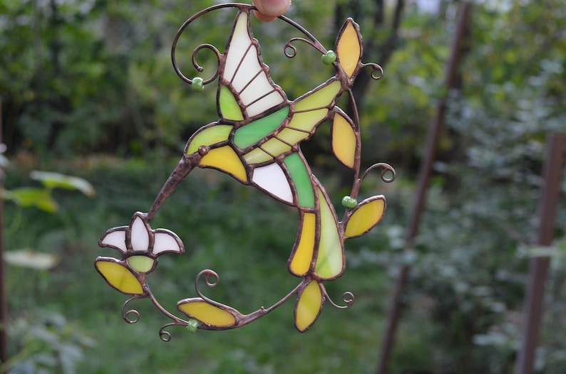 May include: A stained glass hummingbird with green, yellow, and white glass pieces, perched on a branch with a white flower. The hummingbird is in flight with its wings spread.