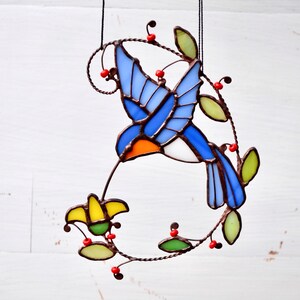 May include: A stained glass hummingbird with blue, orange, and white wings, feeding from a yellow flower. The hummingbird is surrounded by green leaves and red berries.