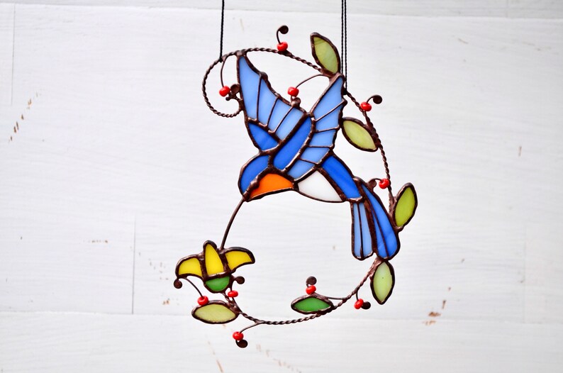 May include: A stained glass hummingbird with blue, orange, and white wings, feeding from a yellow flower. The hummingbird is surrounded by green leaves and red berries.