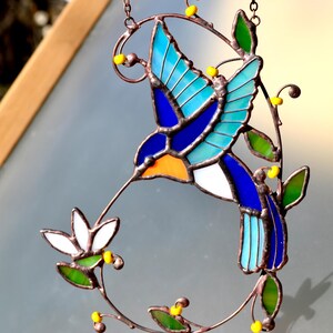 May include: A stained glass hummingbird with blue, green, and orange accents, perched on a copper wire branch with yellow beads. The hummingbird is in flight and has its beak extended as if it is feeding from a flower.